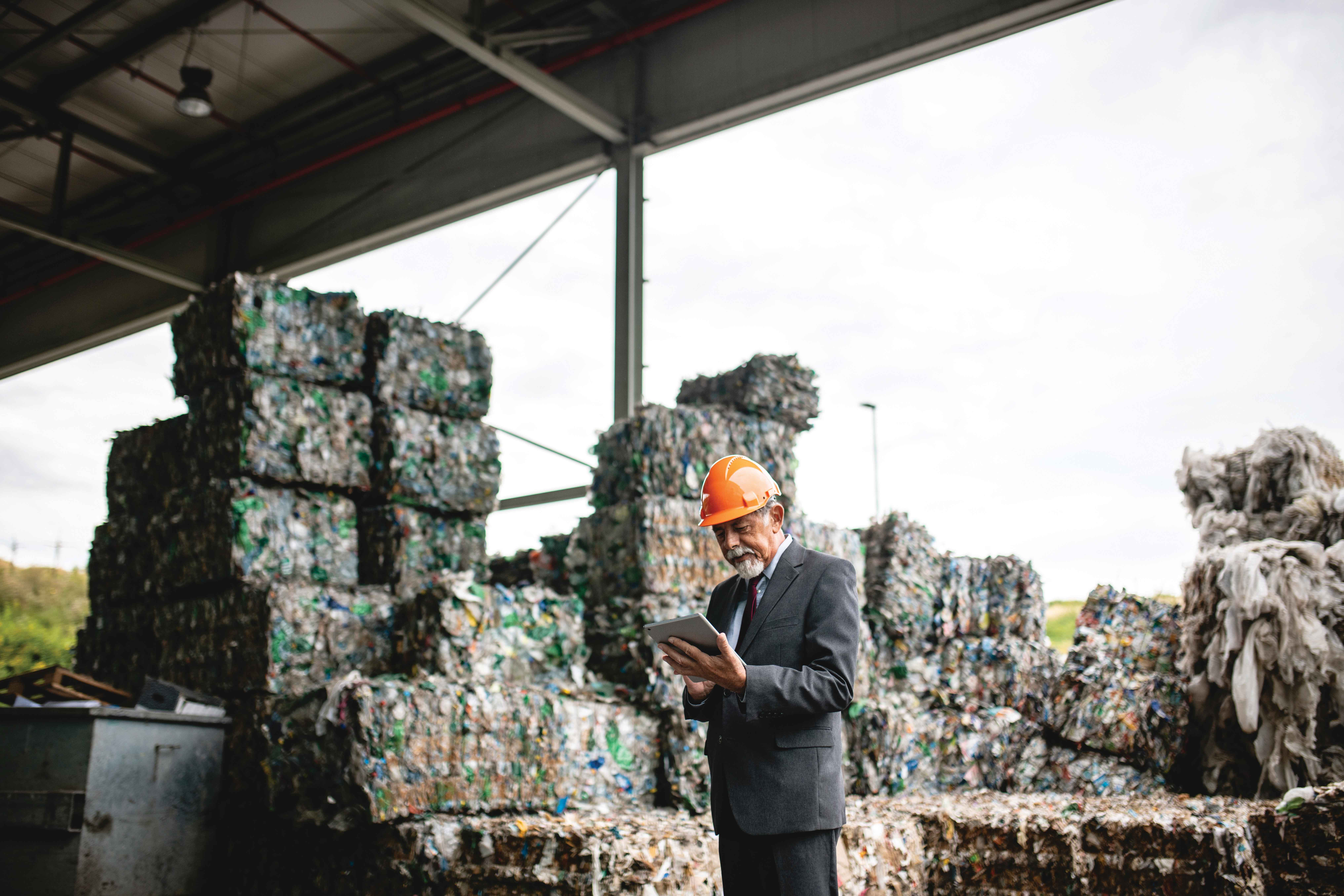 Picture of a businessman with a hard hat on looking at an iPad with packaged waste in the background
